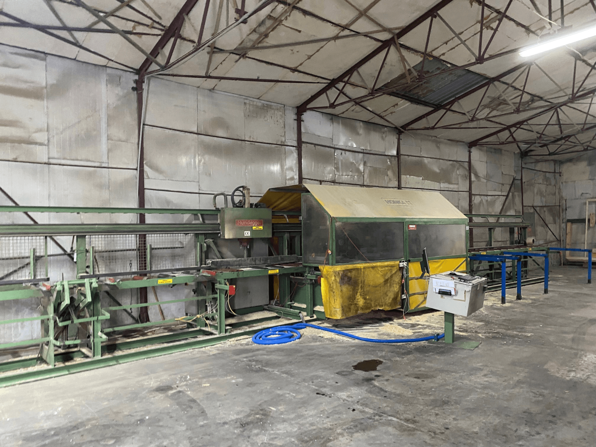 CNC kokapstrādes centrs HUNDEGGER K1 darbnīcā, skats no sāniem, kurā redzamas iekārtas, vadības ierīces un zāģu skaidas uz grīdas.