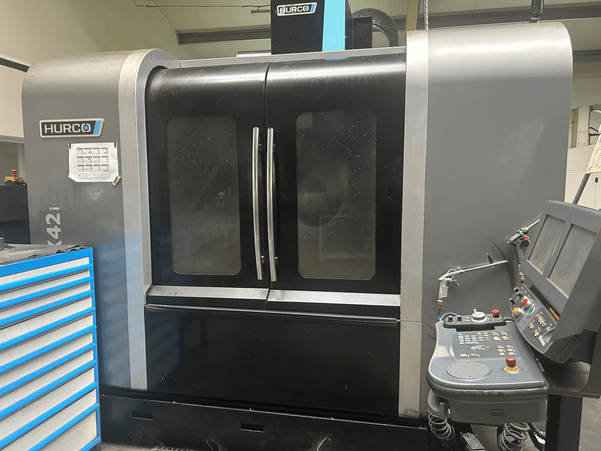 Hurco VMX42i CNC mašīna, skats no priekšpuses ar vadības paneli sānos, ar dubultām durvīm un redzamām tehniskām detaļām.