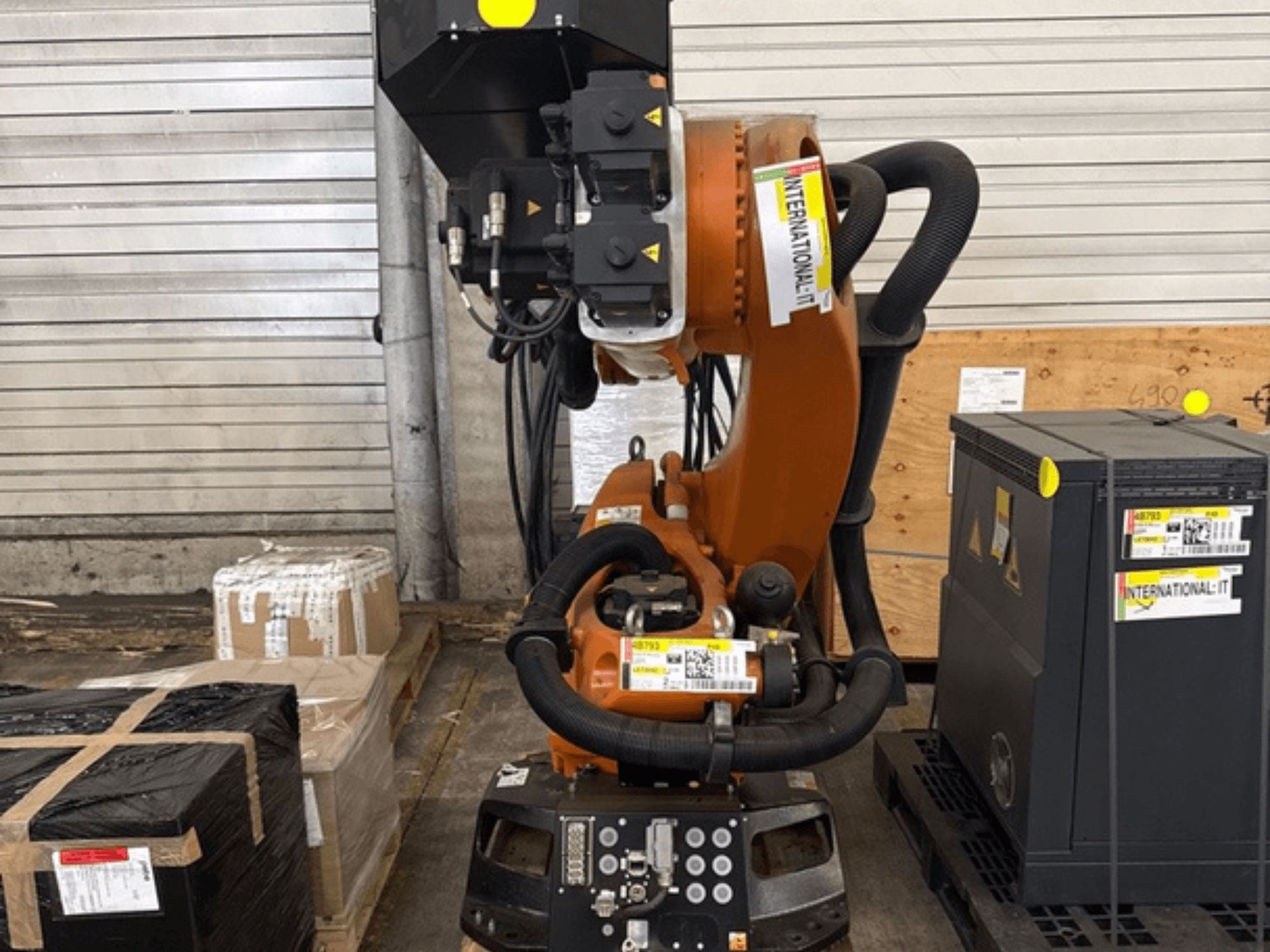 KUKA KR 210 R2700 papildu robota roka, skats no priekšpuses, izstādīta uz paliktņa blakus iepakojumiem un aprīkojumam.