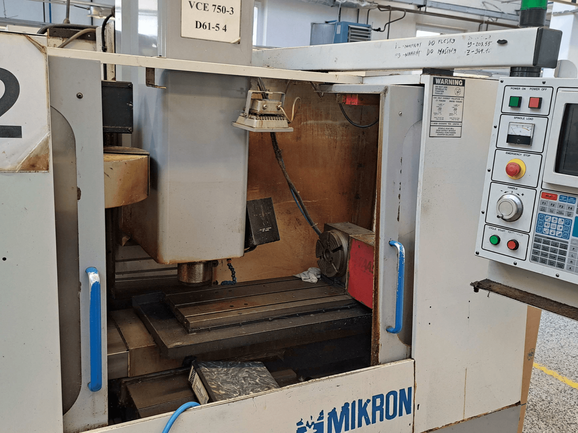 Mikron VCE 750-3 frēzmašīnas priekšējais skats, kurā redzams vārpsta, vadības panelis un darbgalds ar tehniskām detaļām.