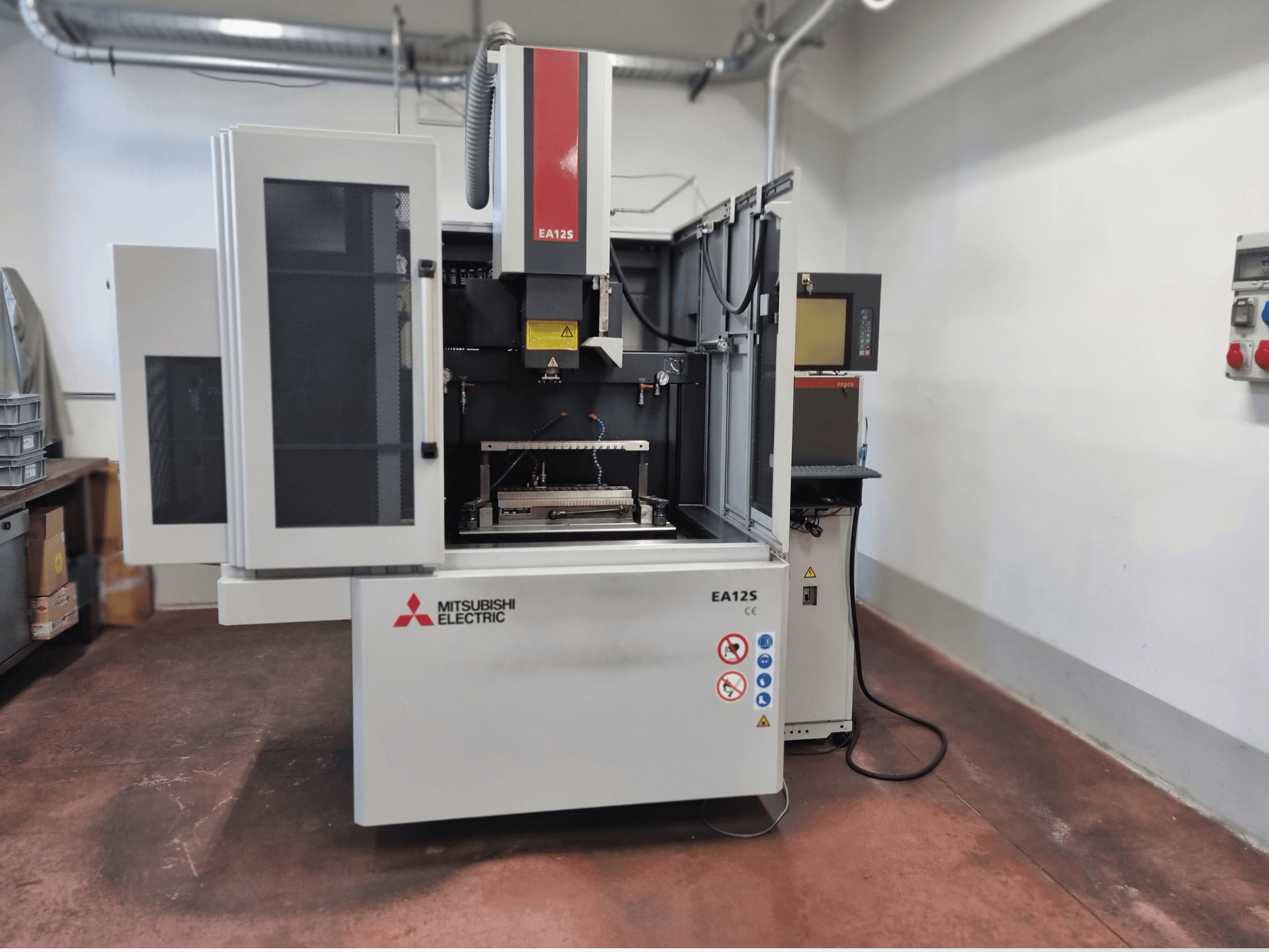 Mitsubishi Electric EA12S stiepļu griešanas iekārta, skats no priekšpuses, ar atvērtām durvīm, vadības paneli un drošības marķējumiem.