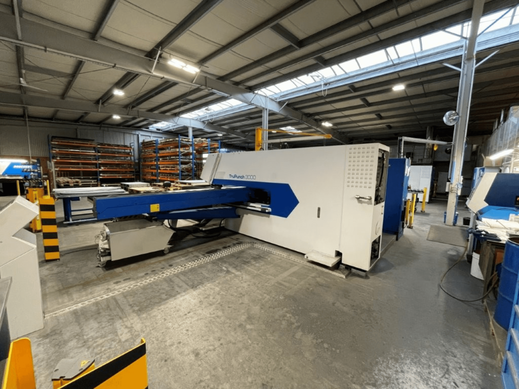 CNC perforācijas preses „TRUMPF TruPunch 3000“ rūpnīcas vidē, skats no priekšpuses, kurā redzams tās zils un balts dizains un darbības komponenti.