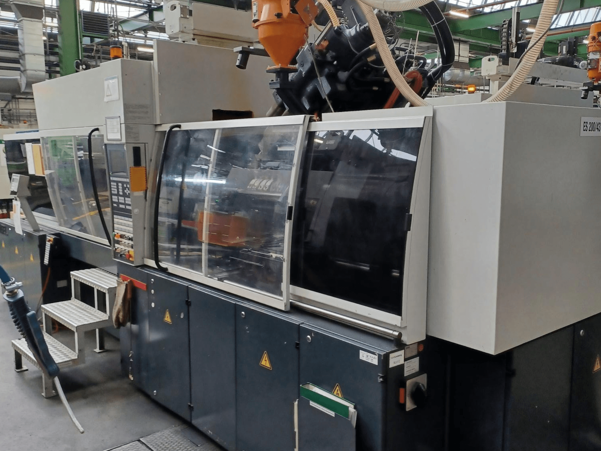 Skats no priekšpuses uz plastmasas iesmidzināšanas formēšanas iekārtu, zīmols un modelis nav zināms, ar caurspīdīgām drošības durvīm un vadības paneli.