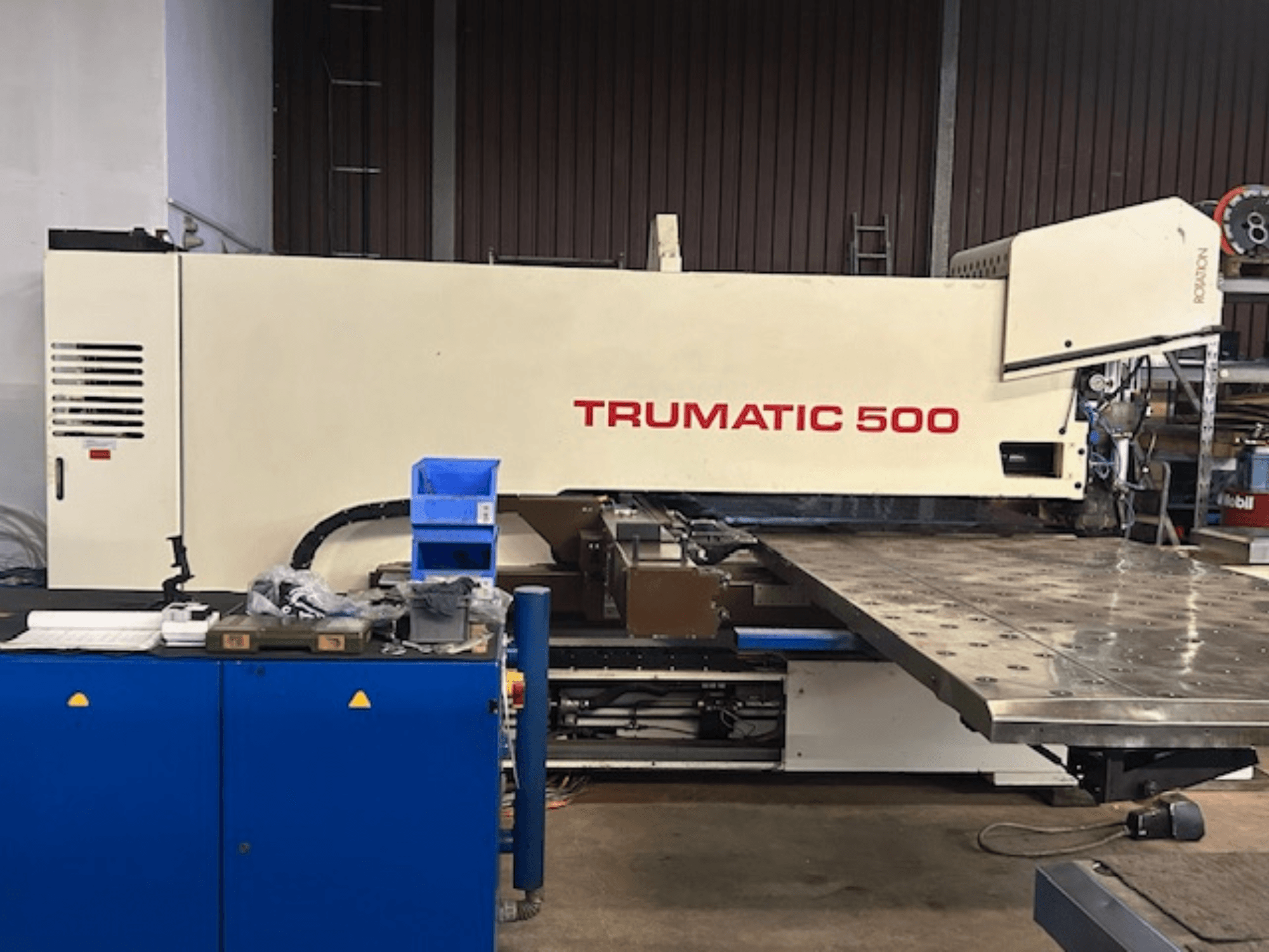 TRUMPF TRUMATIC 500 CNC caurumošanas iekārta, skats no priekšpuses, ar vadības paneli un lielu darba galdu ar perforētu virsmu.
