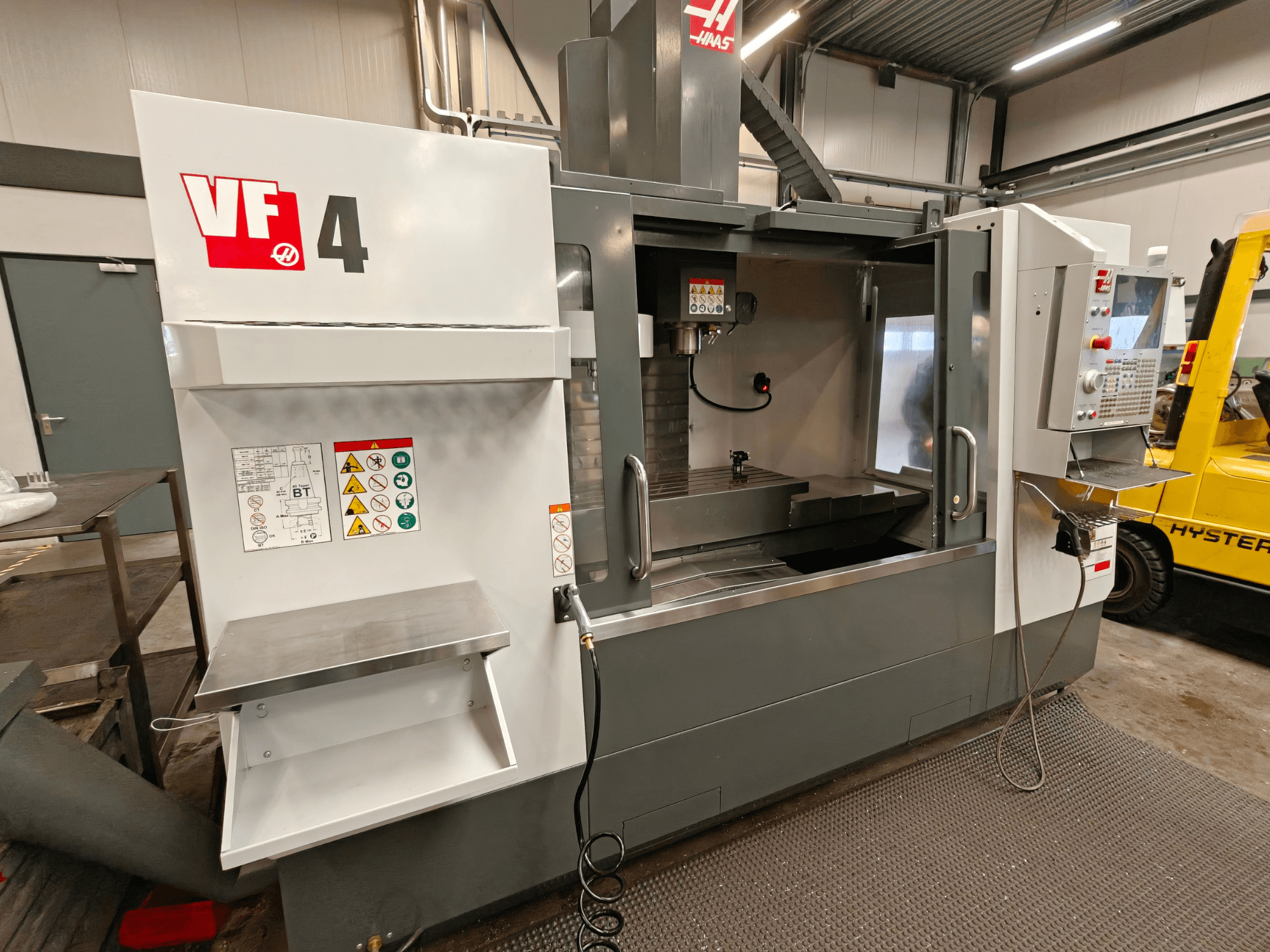 HAAS VF-4 vertikālais apstrādes centrs, skats no priekšpuses, ar vadības paneli, darba zonu un drošības marķējumu sānos.