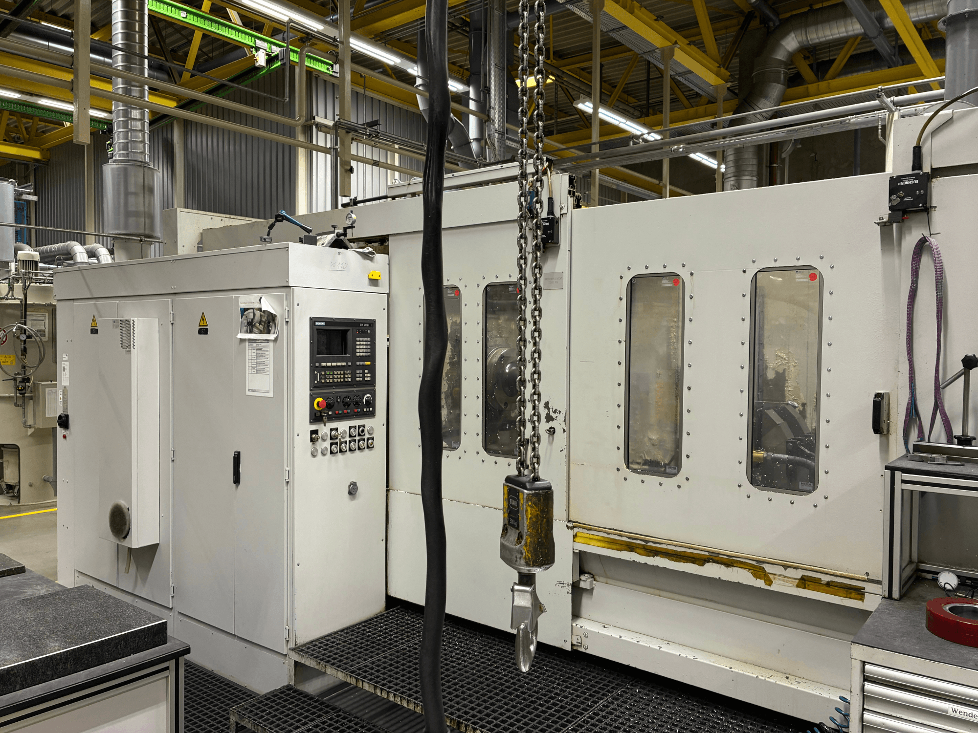 Rūpnieciskā CNC mašīna ar vadības paneli, skats no sāniem; logi, ķēdes un metāla detaļas rūpnīcā.