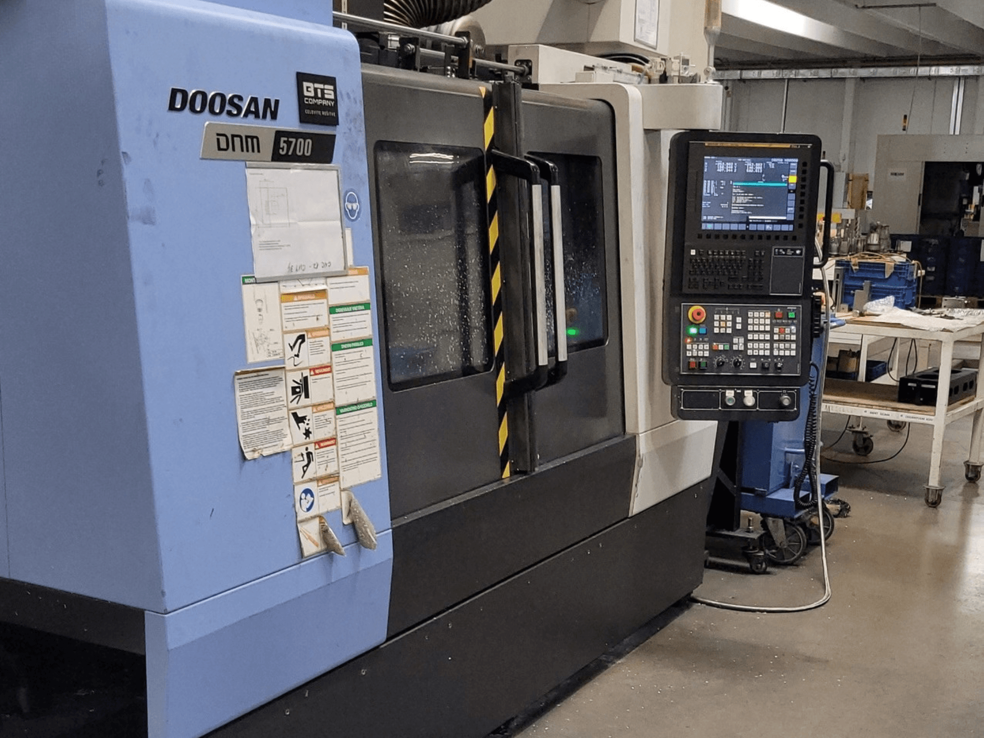 Vertikālais apstrādes centrs DOOSAN DNM 5700, skats no priekšpuses, kurā redzams vadības panelis un darbības vadības elementi.